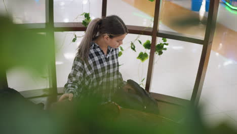 joven se sienta en una silla de madera, desempaquetando artículos de la bolsa mientras que las flores verdes borrosas cubren parcialmente la cara, un ambiente interior acogedor con decoración natural y iluminación suave mejora la atmósfera serena