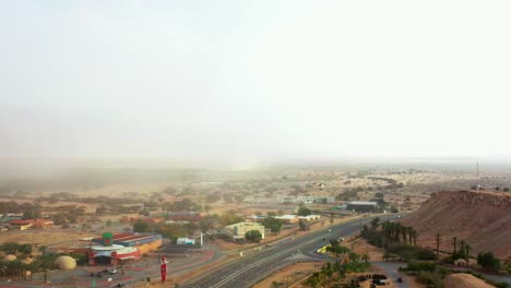 several cars drive on the arabah road highway in the southern negev desert past the large stop shop of the cooperative community kibbutz yotvata with a large sandstorm that rages by