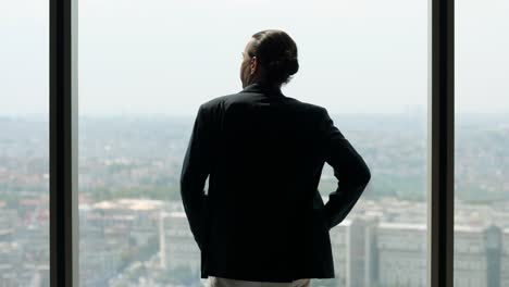 man stands looking out of window at city, taking medical mask off his face