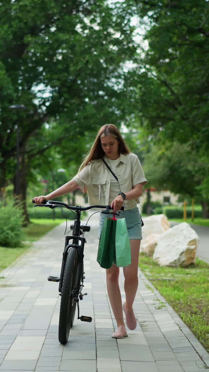 자전거를 타고 산책하는 젊은 성인, 녹색과 민트 가방을 붙인 손잡이를 들고, 평화로운 자연 환경에서 공원 경로를 따라 울창한 초록색, 기둥 및 바위로 둘러싸여 있습니다.