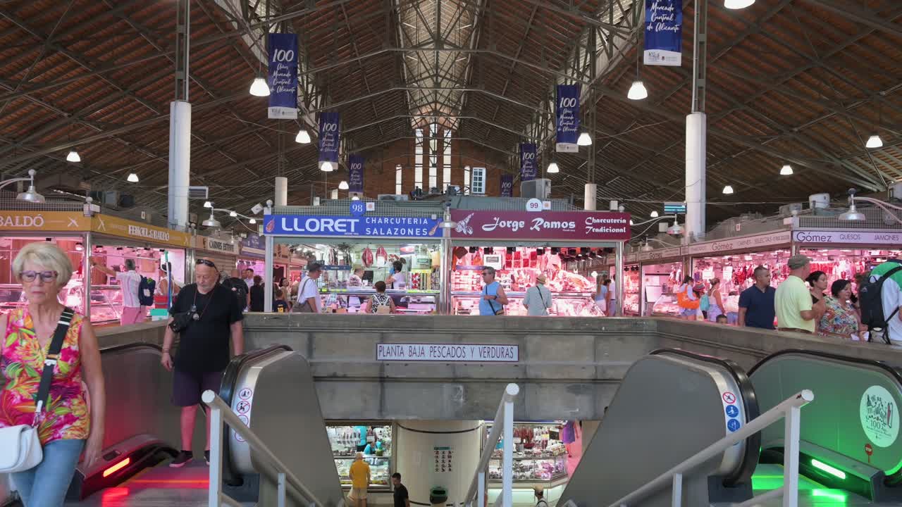 Tilted-down indoor view of Alicante Central Market (built in 1922) as locals shop at various food stalls offering fresh fish, meat, fruits, vegetables, pastries, and tapas in Spain.