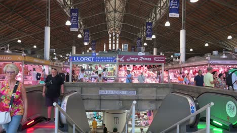 tilted-down indoor view of alicante central market (built in 1922) as locals shop at various food stalls offering fresh fish, meat, fruits, vegetables, pastries, and tapas in spain.