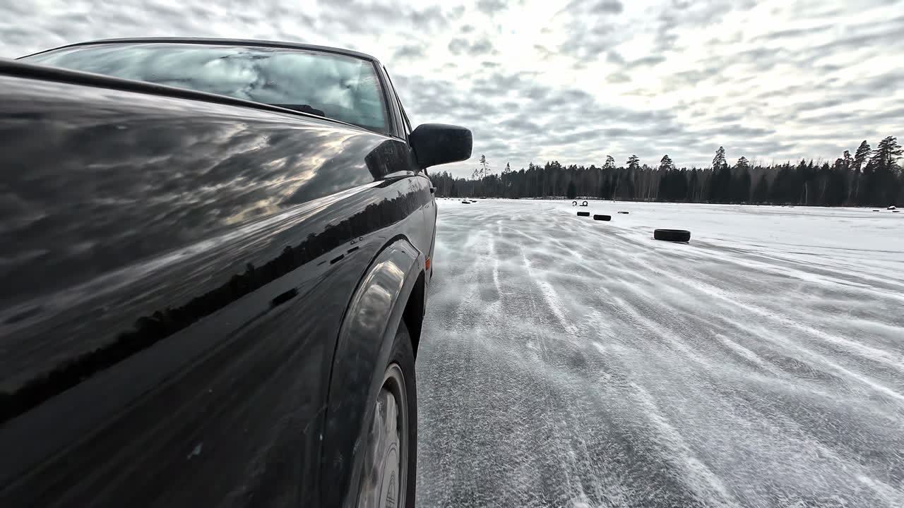 Black car powers through snowy drifts on an icy lake circuit, tire tracks carving curves under cloudy winter skies with forest backdrop