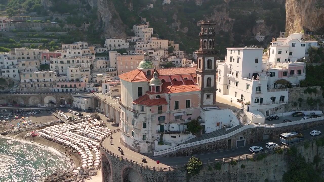 Mostra la Costiera Amalfitana in Italia con edifici colorati e una chiesa vicino al mare. Barche visibili nell'acqua. Il sole splende sulle colline e sul mare.