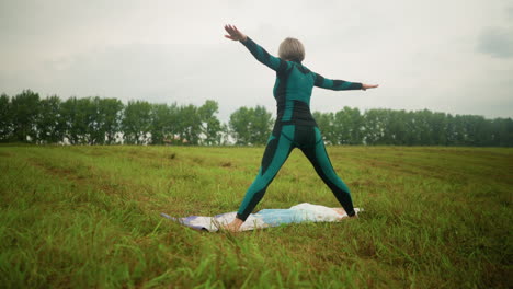 back view of woman in green black suit on yoga mat practicing triangle forward pose, arms extended, standing in open grassy field under cloudy skies with trees and greenery in the background