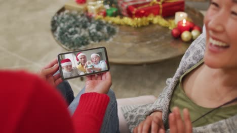 Pareja-Asiática-Sonriente-Usando-Un-Teléfono-Inteligente-Para-Una-Videollamada-Navideña-Con-Una-Familia-Con-Sombreros-De-Papá-Noel-En-La-Pantalla