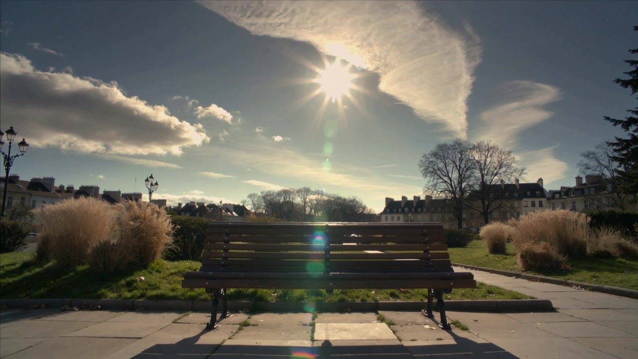 Wanderndes Sonnenlicht wirft den langen Schatten einer Holzbank über einen gepflasterten Platz in einem Stadtpark und markiert die Zeit.