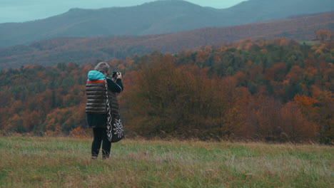 Ältere-Fotografin-Mit-Grauen-Haaren-Fotografiert-Mit-Ihrer-Kamera-In-Der-Natur,-Umgeben-Von-Herbstlich-Gefärbten-Bäumen-Und-Gras,-An-Einem-Kalten,-Windigen-Tag-In-Zeitlupe