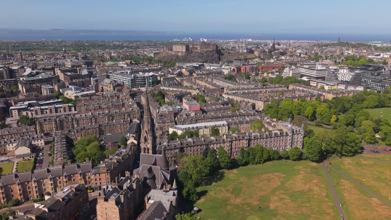Uma vista aérea panorâmica de Edimburgo revela o exuberante parque Meadows com Arthur’s Seat elevando-se ao fundo sob céus azuis brilhantes.