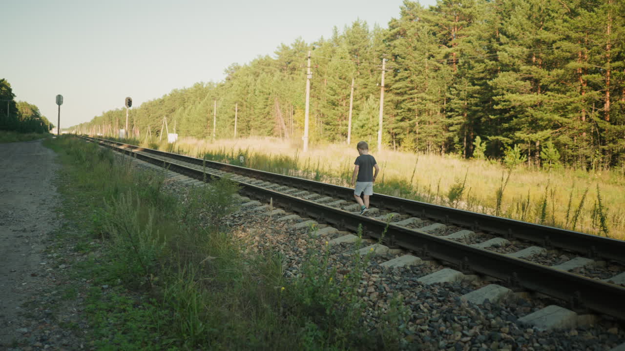 little child carefully walking on railway track surrounded by grassy terrain and utility poles with dense forest background under soft sunlight in peaceful countryside setting