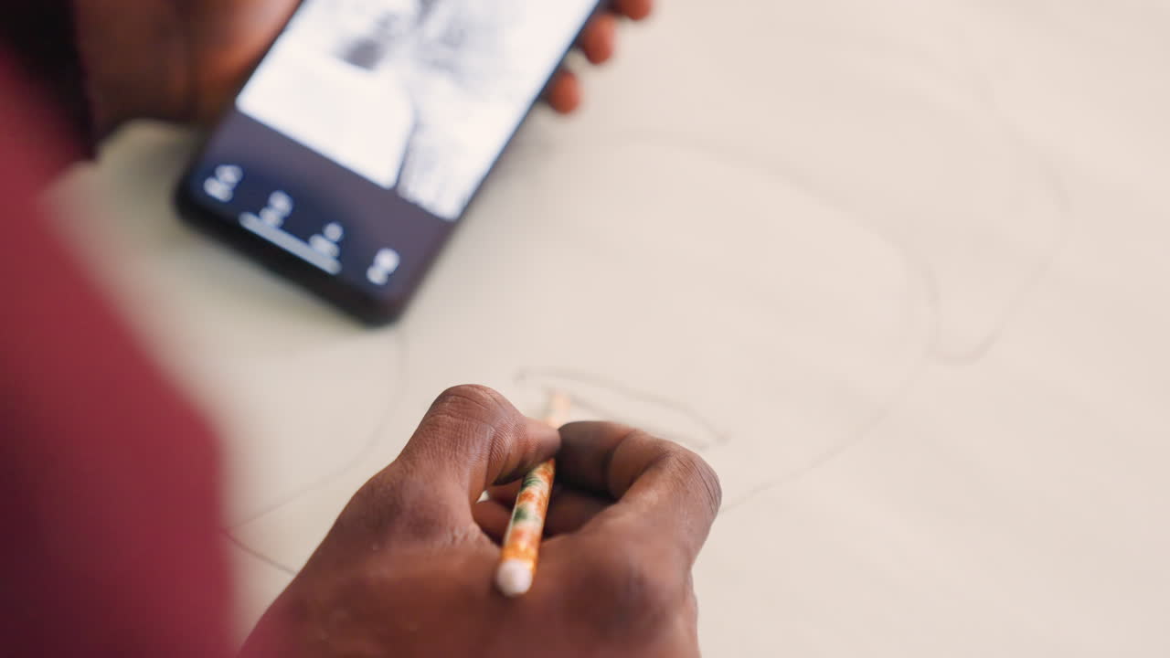 Artista nero che schizza un ritratto da smartphone. Primo piano di mani dalla pelle scura con matita su carta, illuminazione calda e soffusa, ombre delicate, tratti costanti, telefono che mostra l'immagine di riferimento, processo creativo