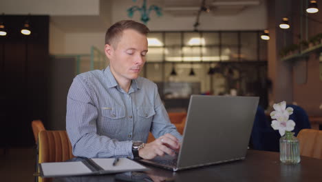 A-young-man-in-a-shirt-is-sitting-at-a-table-with-a-laptop-and-typing-on-the-keyboard.-A-student-can-study-remotely.-A-businessman-conducts-his-business-remotely