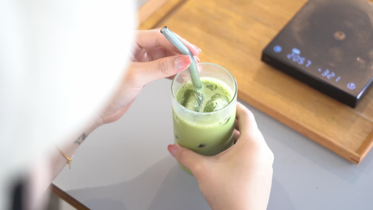 toma cinematográfica sobre el hombro de una joven que bebe una bebida refrescante de verano, un popular té verde matcha helado con leche con un vaso alto y paja