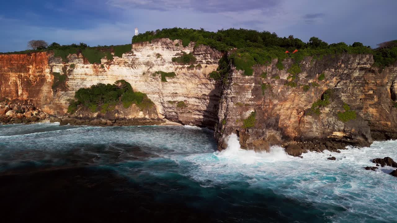 Smooth aerial footage showing a steep Bali cliff with bright rock layers and dense greenery above foamy waves crashing below, highlighting strong ocean motion and striking rugged coastline scenery
