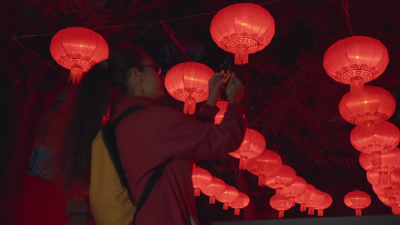 Mulher asiática fotografando lanternas vermelhas à noite, mãos enquadrando a foto com o smartphone, mochila e atmosfera de viagem, brilho carmesim luminoso, cenário de festival cultural, foco em composição criativa, espontânea.