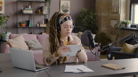 mujer con rastas grabando un podcast hablando en un micrófono sentado en el escritorio con una computadora portátil y documentos