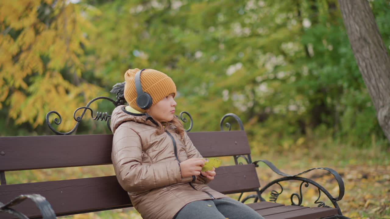 young girl seated on ornate iron bench under autumn canopy wearing mustard beanie and headphones, thoughtfully turning yellow leaf in hand while listening to music amid fallen foliage in park