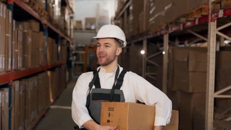 a handsome man in a helmet and work uniform walks in a warehouse with a cardboard box in his hands