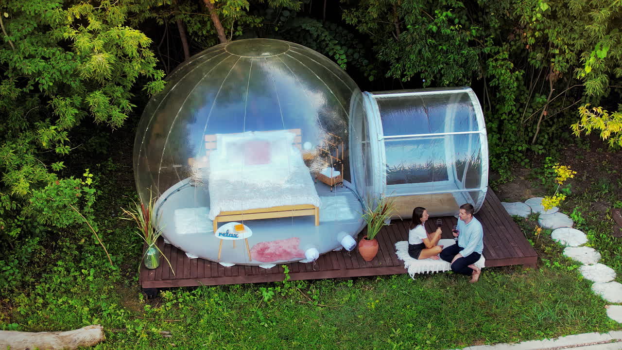 Drone view of a happy couple drinking wine near transparent bubble tent at glamping. Nature around