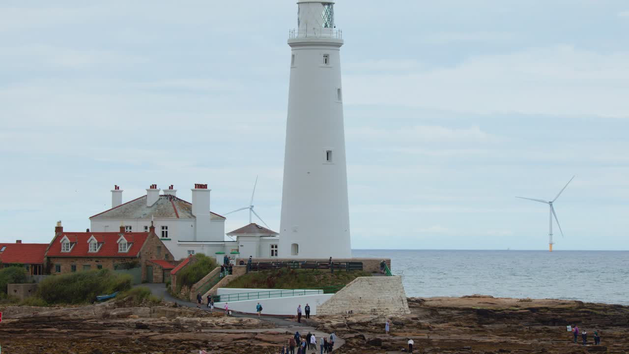 Wide shot of St Mary’s Lighthouse, adjacent buildings, and rocky coastline with people walking. Overcast daylight, static camera, distant wind turbines visible