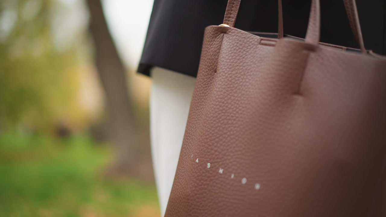Primo piano di una donna che si sistema i lunghi capelli scuri mentre tiene la tracolla di una borsa di pelle marrone, indossando un cappotto nero e una camicia bianca. L'attenzione rimane sui movimenti delle mani con sottili cambiamenti nella presa.