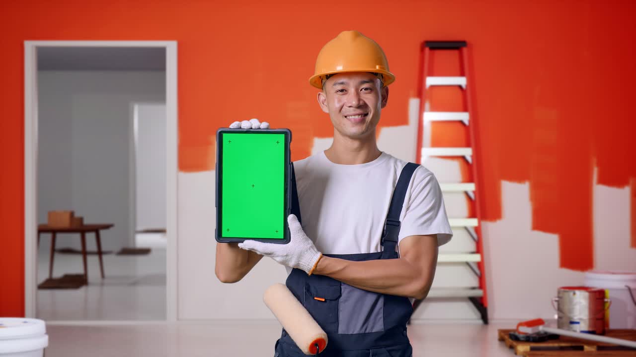 Asian Man Painter Wearing Safety Helmet Smiling And Showing Green Screen Tablet To The Camera While Standing With Painting Wall In Room