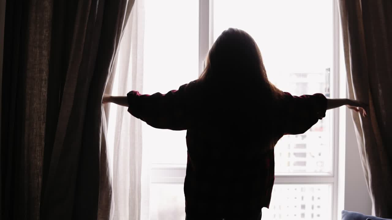 여성의 실루 ⁇ 이 베이지색 커튼을 열고 빛이 방으로 들어옵니다.