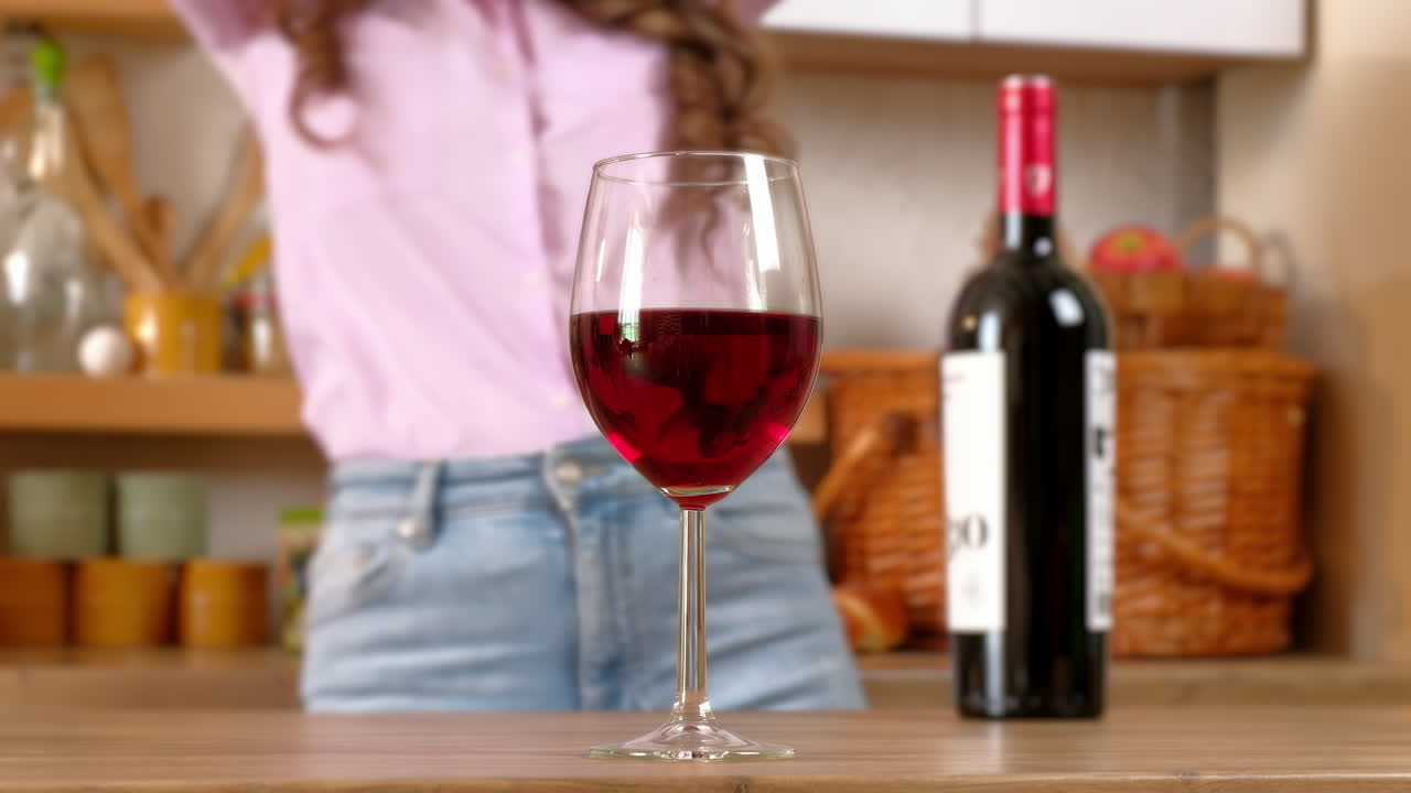 A glass and a bottle of red wine standing on the table in front of a woman dancing in the kitchen