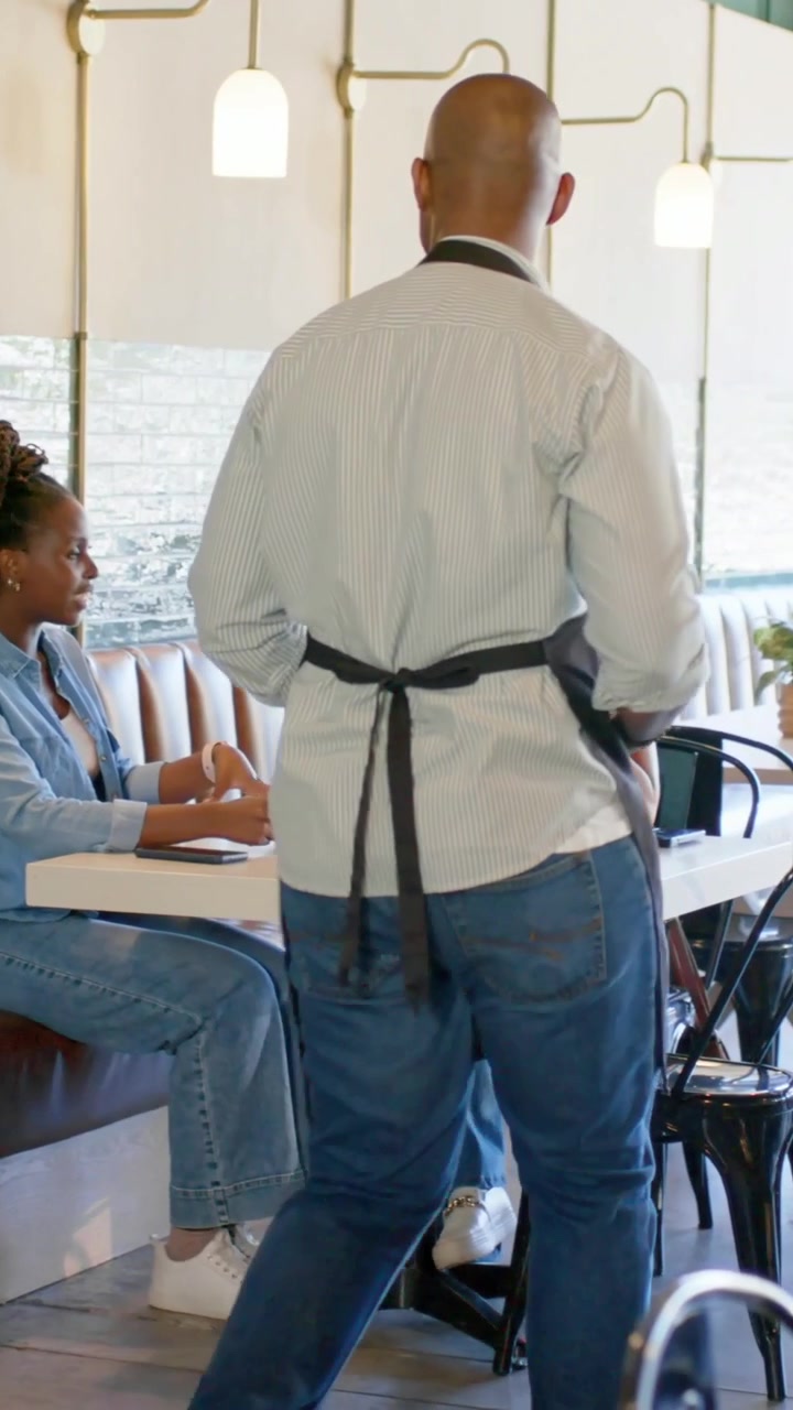 Vertical video: Walking server in light shirt apron carrying pad after alert serving diners in cafe