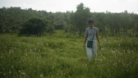 young woman in striped t-shirt and denim jeans walking through tall grass field with trees in the background