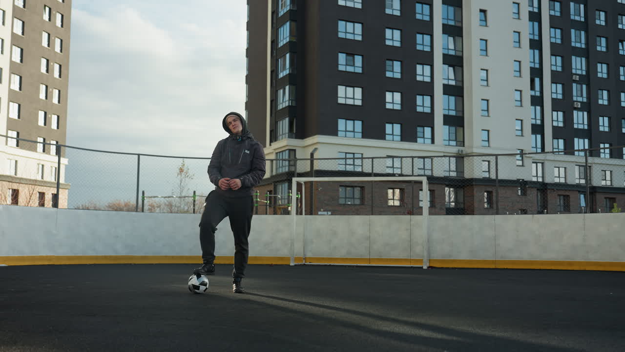 atleta con capucha con la pierna en la pelota estirando los brazos de ancho, girando la cabeza en la arena deportiva urbana, rodeado de postes de gol y altos edificios residenciales