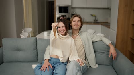 smiling millennial couple home owners sitting on cozy sofa, showing keys to camera, feeling excited