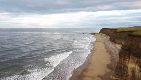 Drohnenaufnahme,-Die-über-Den-Rand-Einer-Grasbewachsenen-Klippe-Fliegt-Und-Am-Abend-Einen-Wunderschönen-Sandstrand-Und-Ruhiges-Meerwasser-Offenbart