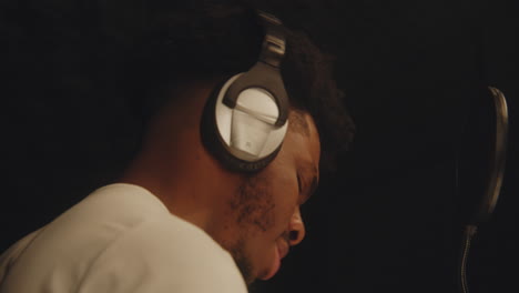 african american singer in headphones dances, sings lyrical composition into microphone in soundproof room and gesticulates to audio engineer. man works in sound recording studio. close up. zoom in.