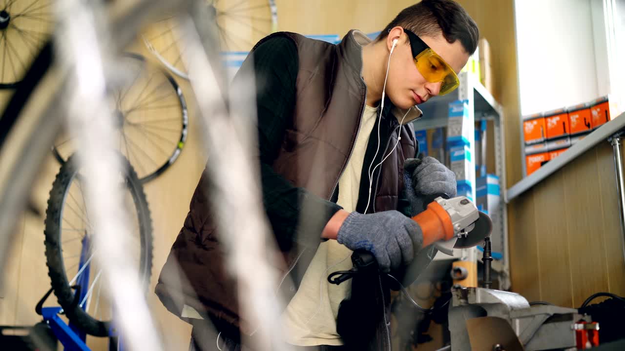Young good-looking man experienced serviceman is using electric circular saw in his workshop. Guy in protective goggles and gloves is looking at saw and listening to music.
