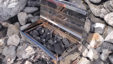 Man-hands-ignite-barbecue-fire-Charcoal-Grill-on-stones-at-beach,-coastal-lifestyle