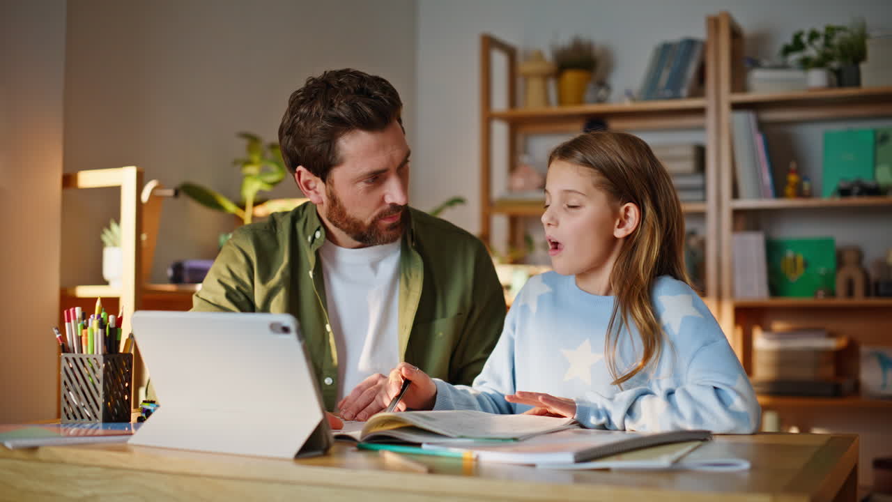 Primo piano di un padre che aiuta la figlia durante una lezione online, seduti insieme a un tavolo in appartamento