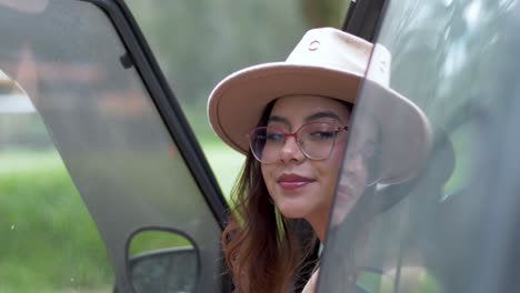 a woman with glasses and hat comes out from car, greeting the camera with a friendly smile and fashionable flair