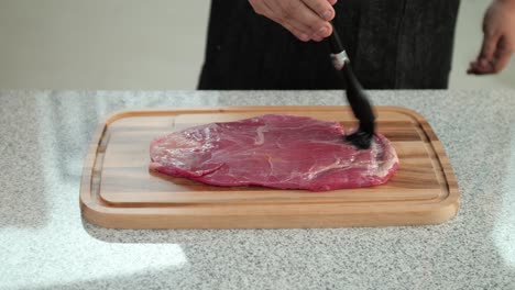 close-up of a black silicone brush smearing beef meat with olive oil on a cutting board