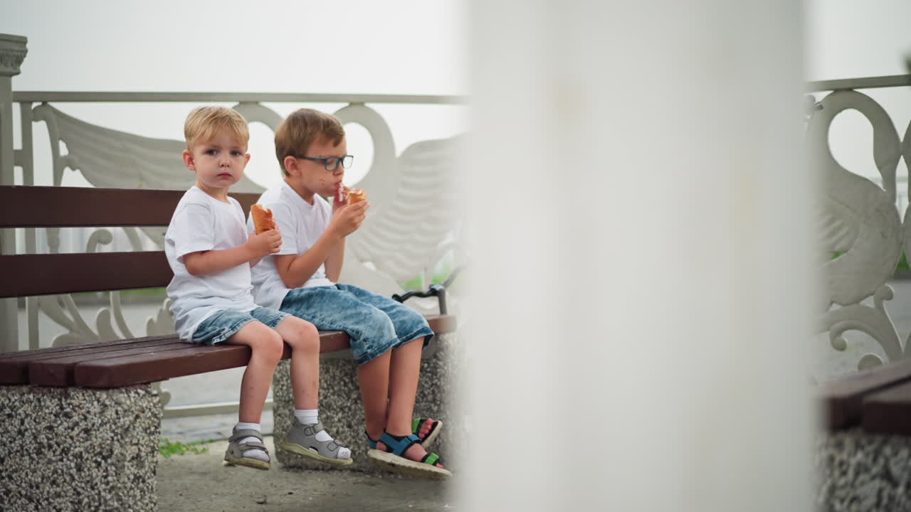Two young siblings sit together on a bench, the older child is eating a sausage roll, while the younger one is holding looking focus, a scooter is near by