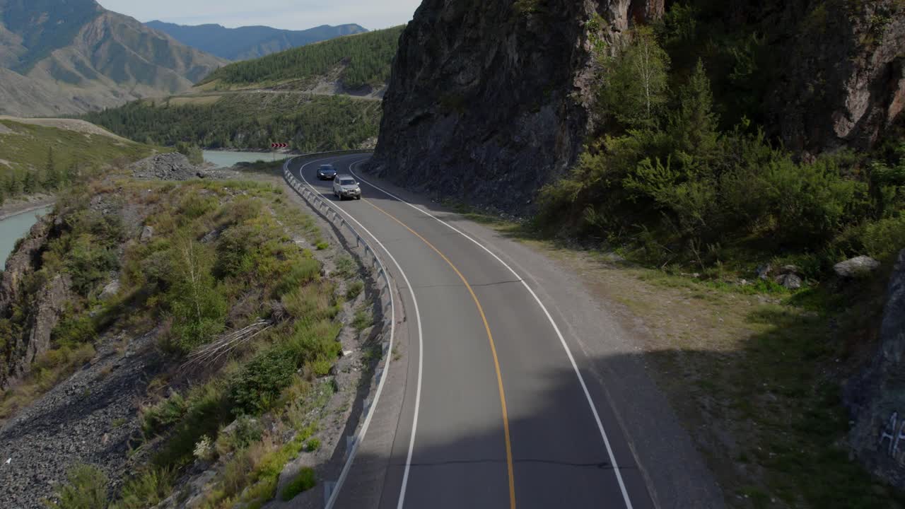 Imágenes de dron de una carretera asfaltada sinuosa que serpentea junto al río Katún turquesa, con acantilados rocosos y montañas boscosas en la República de Altái, Rusia.