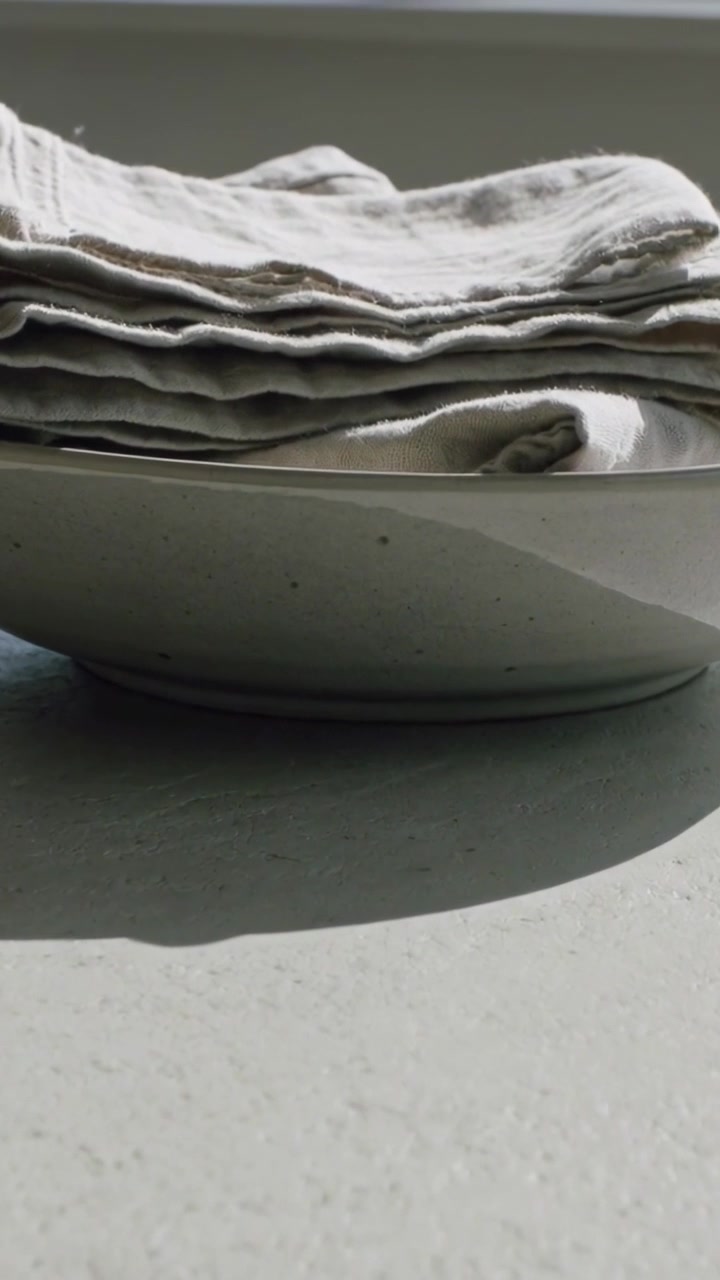 Vertical video: Tilting camera closer to stacked linen napkins in bowl at window, revealing texture