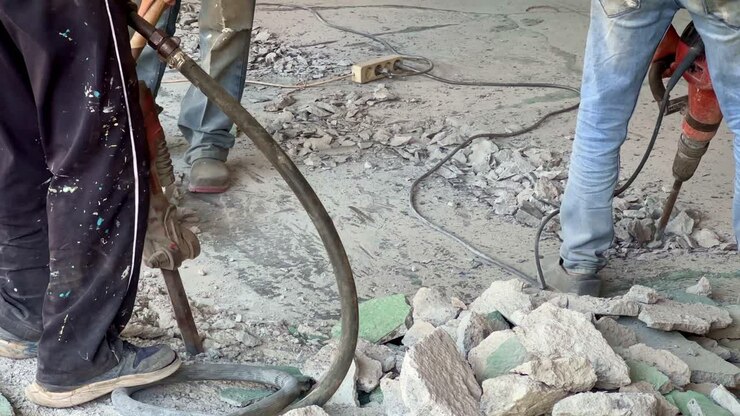 Workers operating jackhammers during concrete floor demolition in an indoor construction site.