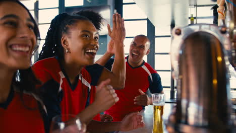 multi-cultural group of friends wearing team shirts in sports bar celebrating watching game on tv