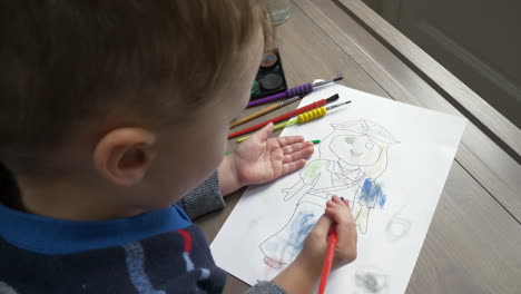 a mid shot of a young boy at a desk, filling brush from a paint set and painting in a pre drawn figure