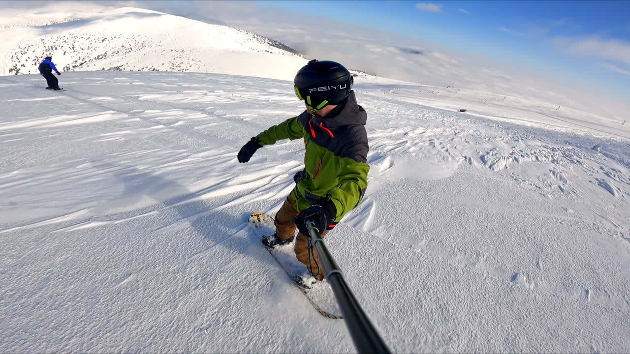 Sportsman with a selfie stick rides down the slope on his snowboard. Winter sport activity.