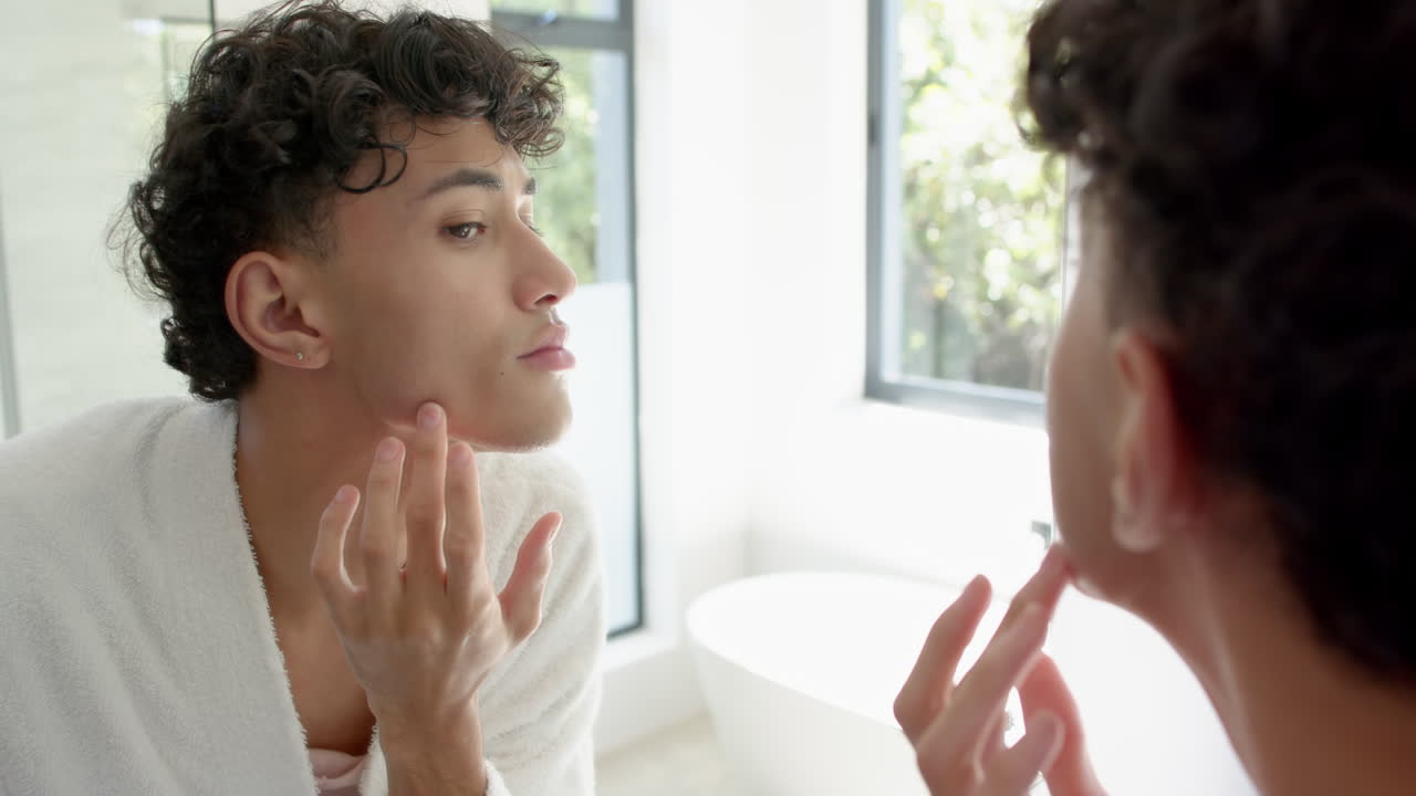 Applying skincare, non binary person examining face in bathroom mirror, focusing on skin