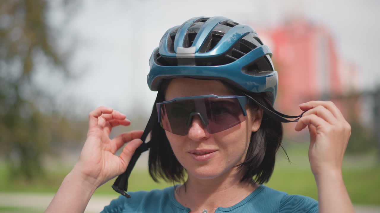 white woman adjusting sunglasses and helmet in urban park, short bob hair, teal shirt, fastening chin strap and checking visor calm focused expression, preparing for solo bicycle ride, safety