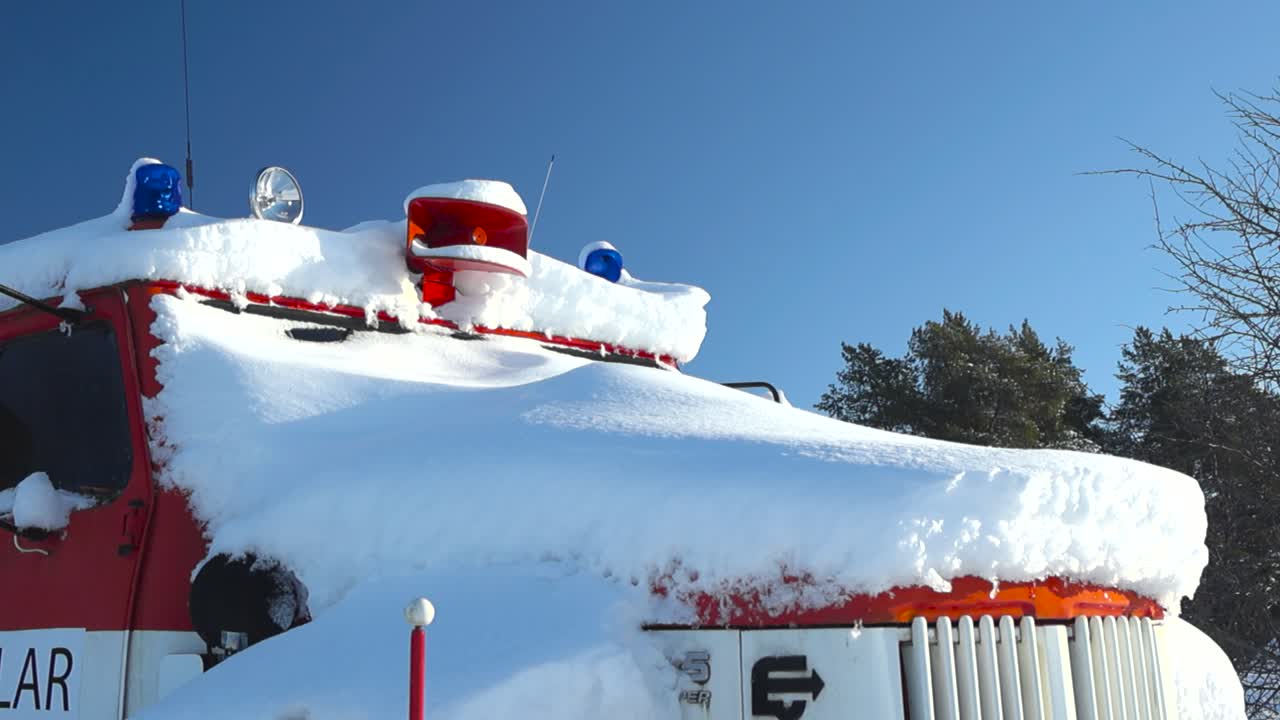 white fluffy snow covered old retro and vintage red firetruck outdoors during a sunny winter day with sun reflecting off the snow. The firetruck has a red trumpet horn siren on roof and flashers also.
