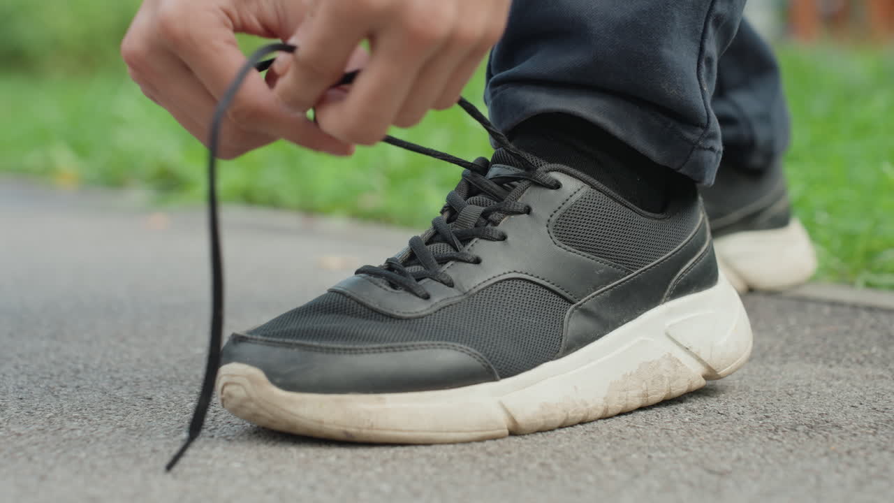 Uomo che si allaccia le scarpe da ginnastica all'aperto, primo piano del processo di allacciatura delle scarpe ai margini di un parco, vista dettagliata di una scarpa da ginnastica casual allacciata vicino al confine erboso del parco durante l'allenamento